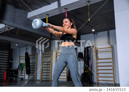 Fitness enthusiast swings a kettlebell in a gym during the day focused on strength training and improving physical health 141635523