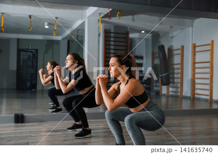 Women perform squats in a gym with wooden floors and exercise equipment during a workout session in the morning hours Women perform squats in a gym with wooden floors and exercise equipment during a workout session in the morning hours 141635740