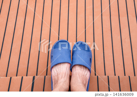 Top down view of feet wearing blue rubber sandals on a teak deck with black lines. Relaxing vacation vibe on a boat or luxury yacht. Concept of leisure, summer travel, and outdoor relaxation. 141636009