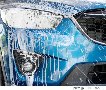 Blue car wash with white soap foam. Auto care business. Car cleaning service. Vehicle cleaning service. Foam wash car detailing. Headlamp light of the blue luxury SUV car is covered with white foam. 141636539