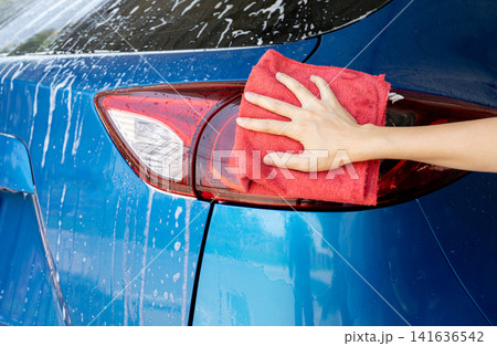 Car wash service. Car covered with white soap foam. Man hand holding red microfiber cloth and polish tail light of blue car. Auto care service business concept. Man cleaning and detailing luxury car. 141636542