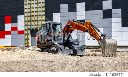 Heavy machinery digs into the ground at a construction site beside a building with bright colors. Workers may operate equipment during the day Heavy machinery digs into the ground at a construction site beside a building with bright colors. Workers may operate equipment during the day 141636574