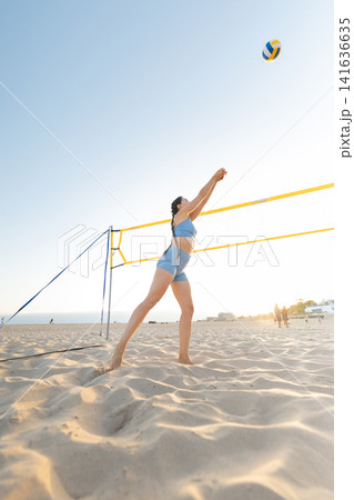 Person plays beach volleyball at sunset during a warm day by the ocean with a volleyball net and sand Person plays beach volleyball at sunset during a warm day by the ocean with a volleyball net and sand 141636635