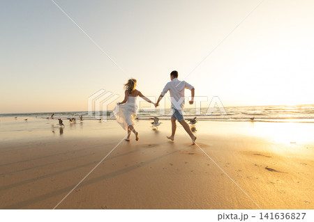 Couple runs along the beach hand in hand as the sun sets in the background with waves crashing and birds flying nearby Couple runs along the beach hand in hand as the sun sets in the background with waves crashing and birds flying nearby 141636827