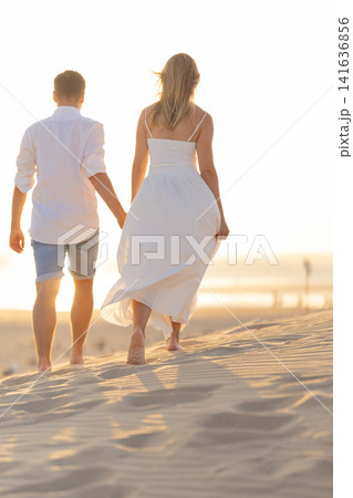 Young couple walks hand in hand on beach sand at sunset near the ocean waves Young couple walks hand in hand on beach sand at sunset near the ocean waves 141636856
