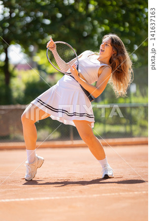 Tennis player takes a swing on a clay court during a sunny day while wearing a white dress and showing determination Tennis player takes a swing on a clay court during a sunny day while wearing a white dress and showing determination 141637163