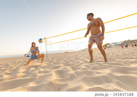 Friends play volleyball on the beach during sunset while other people enjoy the warm sand and clear sky in the background Friends play volleyball on the beach during sunset while other people enjoy the warm sand and clear sky in the background 141637188