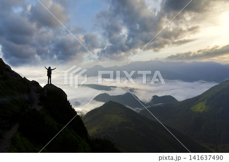Wide mountain panorama. Small silhouette of tourist with backpack on rocky mountain slope with raised hands over valley covered with white puffy clouds. Beauty of nature, tourism and traveling concept Wide mountain panorama. Small silhouette of tourist with backpack on rocky mountain slope with raised hands over valley covered with white puffy clouds. Beauty of nature, tourism and traveling concept 141637490