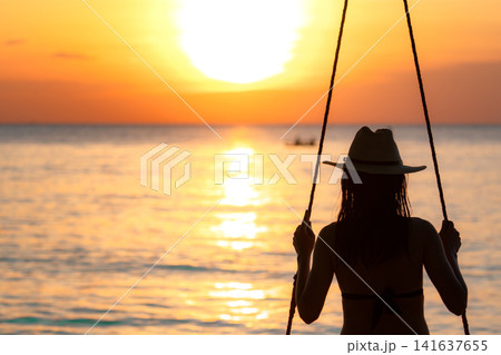Back view of woman on wooden beach swing watching glowing tropical sunset over the calm ocean. Luxury summer vacation, peaceful resort lifestyle, coastal travel and seaside relaxation. Luxury tourism. Back view of woman on wooden beach swing watching glowing tropical sunset over the calm ocean. Luxury summer vacation, peaceful resort lifestyle, coastal travel and seaside relaxation. Luxury tourism. 141637655