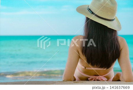 Rear view of woman wearing a straw hat looking at the turquoise tropical ocean, peaceful summer vacation, luxury beach resort, wellness travel destination and relaxation concept. Tourism promotion. Rear view of woman wearing a straw hat looking at the turquoise tropical ocean, peaceful summer vacation, luxury beach resort, wellness travel destination and relaxation concept. Tourism promotion. 141637669