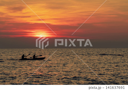 Silhouette of two people kayaking across tropical ocean during a colorful sunset with glowing orange skies. Peaceful adventure travel, outdoor recreation, coastal tourism and summer vacation lifestyle Silhouette of two people kayaking across tropical ocean during a colorful sunset with glowing orange skies. Peaceful adventure travel, outdoor recreation, coastal tourism and summer vacation lifestyle 141637691