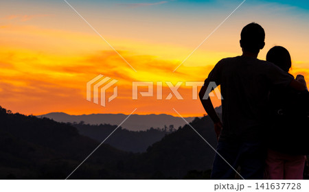 Romantic couple embracing while watching colorful sunset over tropical mountain landscape in Thailand, travel destination with scenic valley, outdoor adventure, tourism and relationship concept. Romantic couple embracing while watching colorful sunset over tropical mountain landscape in Thailand, travel destination with scenic valley, outdoor adventure, tourism and relationship concept. 141637728