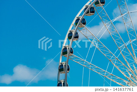 Modern Ferris wheel against blue sky, amusement park attraction for tourism, summer holiday, family entertainment. Amusement ride for travel destination, vacation and funfair entertainment. Modern Ferris wheel against blue sky, amusement park attraction for tourism, summer holiday, family entertainment. Amusement ride for travel destination, vacation and funfair entertainment. 141637761