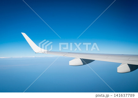 Airplane wing above blue sky and distant clouds viewed from passenger window during an international commercial airline flight. Aviation, travel, transportation and global tourism. Aviation industry, Airplane wing above blue sky and distant clouds viewed from passenger window during an international commercial airline flight. Aviation, travel, transportation and global tourism. Aviation industry, 141637789