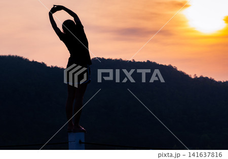 Silhouette of woman stretching at sunrise over mountain landscape during peaceful morning for healthy lifestyle, wellness, mindfulness and outdoor fitness routine. Morning exercise, relaxation. Silhouette of woman stretching at sunrise over mountain landscape during peaceful morning for healthy lifestyle, wellness, mindfulness and outdoor fitness routine. Morning exercise, relaxation. 141637816
