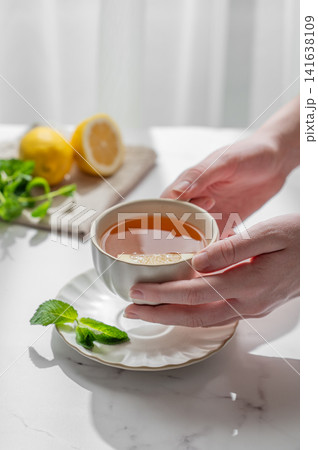 Hand holding a cup of tea with lemon, ginger and mint on a marble background Hand holding a cup of tea with lemon, ginger and mint on a marble background 141638109