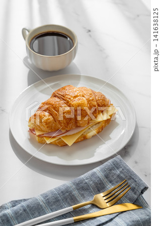 A golden croissant sandwich with ham and cheese on a white plate on a marble background 141638125