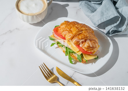 A golden croissant sandwich with ham, cheese, tomato, and arugula on a marble background A golden croissant sandwich with ham, cheese, tomato, and arugula on a marble background 141638132