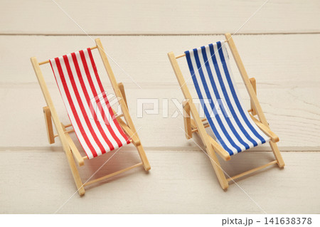 Two miniature deck chairs with red and blue striped canvas seats on light background. Summer concept. Two miniature deck chairs with red and blue striped canvas seats on light background. Summer concept. 141638378