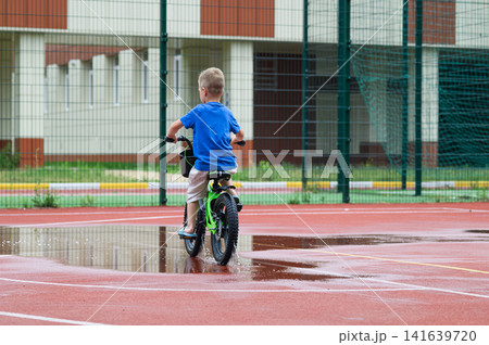Children riding balance bikes through puddles on wet outdoor sports court after rain. Outdoor lifestyle, childhood activity, freedom and play in urban environment. Children riding balance bikes through puddles on wet outdoor sports court after rain. Outdoor lifestyle, childhood activity, freedom and play in urban environment. 141639720