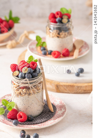 A vertical composition of two glass jars filled with creamy overnight oats and chia seeds layered with granola and fresh berries served on decorative plates with wooden spoons in a bright kitchen A vertical composition of two glass jars filled with creamy overnight oats and chia seeds layered with granola and fresh berries served on decorative plates with wooden spoons in a bright kitchen 141641682
