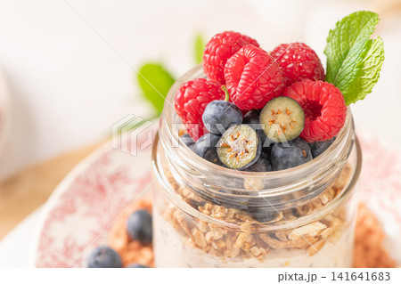 A vibrant horizontal shot of a glass mason jar filled with creamy chia seed overnight oats and granola, topped with an abundance of fresh raspberries and blueberries, garnished with a mint sprig in a 141641683