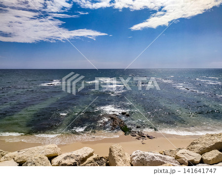 A serene, sunny beach showcasing clear blue ocean waves, sandy shore, and rocky foreground under a vibrant sky. Perfect for inspiring travel and mindfulness designs. No people. A serene, sunny beach showcasing clear blue ocean waves, sandy shore, and rocky foreground under a vibrant sky. Perfect for inspiring travel and mindfulness designs. No people. 141641747