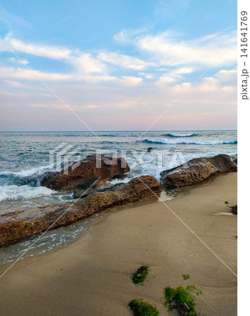 A serene sandy beach features mossy rocks meeting gentle ocean waves under a beautiful pastel sunset sky. This peaceful coastal landscape with no people evokes a sense of deep calm. A serene sandy beach features mossy rocks meeting gentle ocean waves under a beautiful pastel sunset sky. This peaceful coastal landscape with no people evokes a sense of deep calm. 141641769