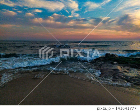 Serene ocean waves crash onto a sandy shore at sunset, showcasing a dramatic sky. This tranquil seascape evokes peace and mindfulness. Perfect for travel and meditation. Nobody. 141641777