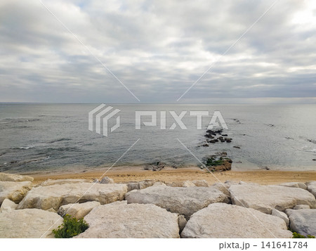 A serene, contemplative view of a quiet ocean beach under a cloudy sky. Nobody present. The rocky foreground evokes a sense of natural stability and peace, ideal for travel or coastal themes. A serene, contemplative view of a quiet ocean beach under a cloudy sky. Nobody present. The rocky foreground evokes a sense of natural stability and peace, ideal for travel or coastal themes. 141641784