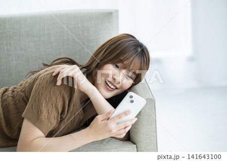 ソファに寝転がってスマホを使う若い女性 141643100