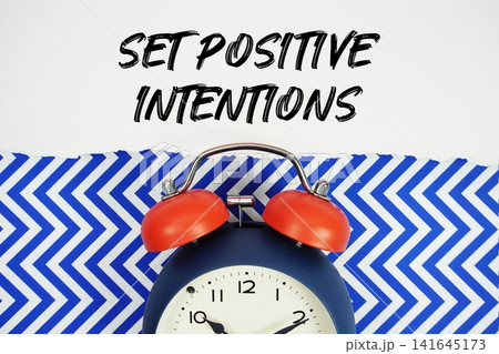 Set Positive Intentions text written on paper with alarm clock on blue chevron background Set Positive Intentions text written on paper with alarm clock on blue chevron background 141645173