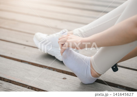 Woman stretching foot after running with muscle pain and leg injury, female athlete sits on wooden boardwalk holding painful foot in white sock during workout recovery outdoors 141645627