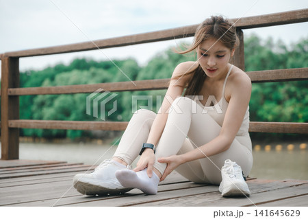 Woman runner athlete holding ankle, suffering muscle tendon pain, feeling intense sport injury discomfort while sitting on wooden bridge outdoor, needing medical treatment Woman runner athlete holding ankle, suffering muscle tendon pain, feeling intense sport injury discomfort while sitting on wooden bridge outdoor, needing medical treatment 141645629