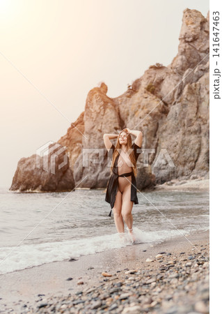 Woman beach swimwear walking along the pebble shore with rocky cliffs at sunset enjoying the calm sea waves Woman beach swimwear walking along the pebble shore with rocky cliffs at sunset enjoying the calm sea waves 141647463