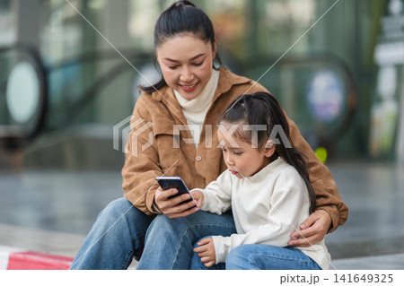 mother and daughter sitting down, happy and using smartphone in city 141649325