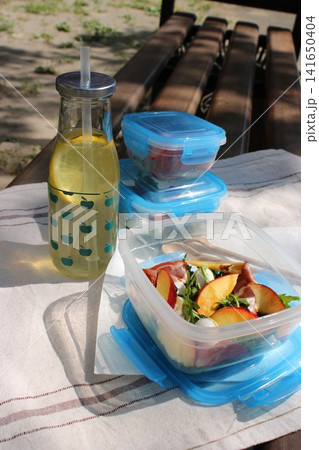 Clear container with salad and fruits placed on a picnic table outdoors, accompanied by a bottle of lemonade, with greenery in the background Clear container with salad and fruits placed on a picnic table outdoors, accompanied by a bottle of lemonade, with greenery in the background 141650404