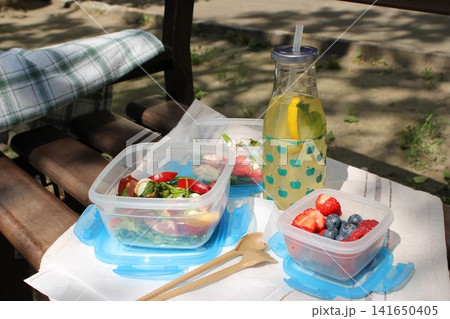 Fresh salad and mixed berries in clear containers with a bottle of lemonade on a picnic table, surrounded by greenery in a sunny outdoor setting Fresh salad and mixed berries in clear containers with a bottle of lemonade on a picnic table, surrounded by greenery in a sunny outdoor setting 141650405