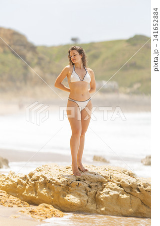 Female model in white bikini poses on a rock by the ocean, with waves crashing and green hills visible in the background under a clear sky Female model in white bikini poses on a rock by the ocean, with waves crashing and green hills visible in the background under a clear sky 141652884