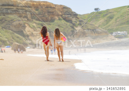 Two women in swimsuits walking along a sandy beach, one in red and the other in white, with cliffs and ocean waves in the background on a sunny day 141652891