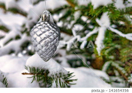 A silver pinecone ornament hangs from a decorated tree covered in snow during the Christmas season. The scene shows a typical winter landscape A silver pinecone ornament hangs from a decorated tree covered in snow during the Christmas season. The scene shows a typical winter landscape 141654356