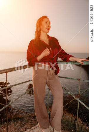 Woman sunset ocean standing on cliffside balcony enjoying coastal view during golden hour Woman sunset ocean standing on cliffside balcony enjoying coastal view during golden hour 141654622