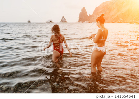 Women beach ocean, two friends walking into the sea from a rocky shore during sunset. Women beach ocean, two friends walking into the sea from a rocky shore during sunset. 141654641