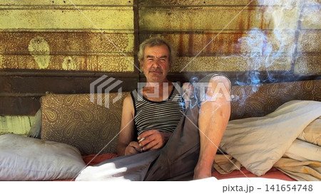Portrait of homeless adult man sitting on worn couch placed and smoking outdoor. The setting underscores neglect and impermanence, presenting a raw and unfiltered glimpse of difficult living situation 141654748
