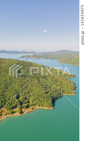 Vertical aerial view of turquoise mountain lake coast with green forest peninsula and distant hills in Plastiras Thessaly Greece for travel destination background Vertical aerial view of turquoise mountain lake coast with green forest peninsula and distant hills in Plastiras Thessaly Greece for travel destination background 141654975