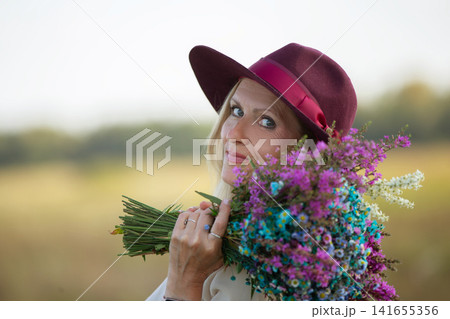 Woman in a burgundy hat holding a colorful bouquet of wildflowers in a sunny field during daytime Woman in a burgundy hat holding a colorful bouquet of wildflowers in a sunny field during daytime 141655356