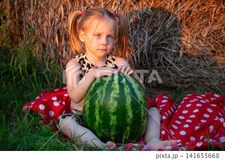Little girl enjoys summer day with large watermelon while sitting on colorful blanket near hay bales Little girl enjoys summer day with large watermelon while sitting on colorful blanket near hay bales 141655668