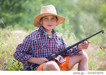 A cheerful boy dressed in a checkered shirt and a straw hat sits with a fishing rod, smiling at the camera. The setting features a vibrant green riverbank and bright sunlight. 141655670