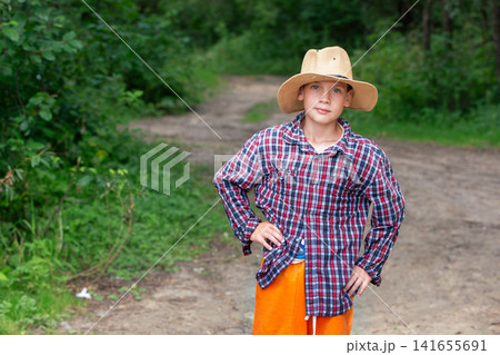 Young boy in a straw hat stands proudly on a forest path surrounded by greenery during daytime 141655691