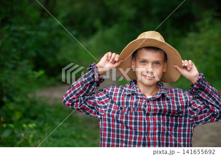 A young boy stands confidently in a plaid shirt and straw hat. He is smiling while posing in a lush green environment, enjoying a sunny day outdoors. A young boy stands confidently in a plaid shirt and straw hat. He is smiling while posing in a lush green environment, enjoying a sunny day outdoors. 141655692